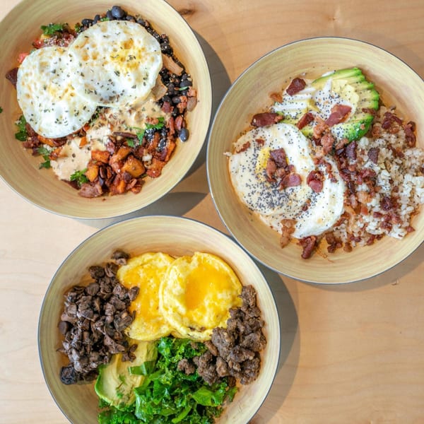 Three healthy breakfast bowls with eggs, avocado, rice, beans, and various toppings on a wooden table.