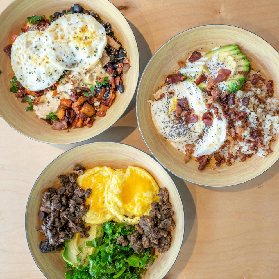 Three healthy breakfast bowls with eggs, avocado, rice, beans, and various toppings on a wooden table.