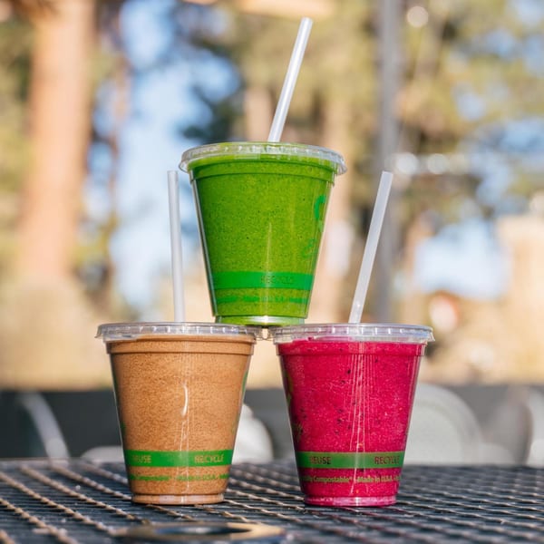 Three colorful smoothies from Life & Time, green, brown, and red, stacked on an outdoor table.