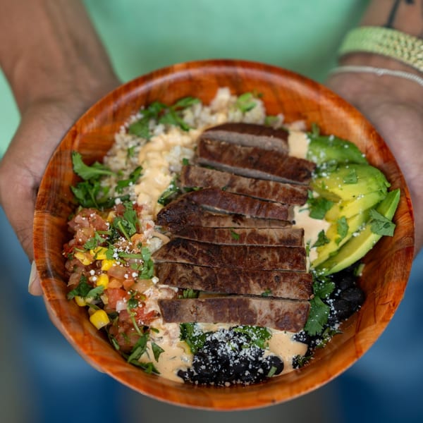 Hands holding a wooden bowl filled with sliced steak, avocado, salsa, black beans, and rice.