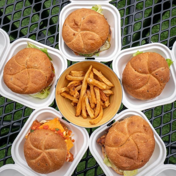 Five Life & Time burgers in takeout containers surround a bowl of fries on a black mesh table.