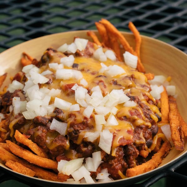 Sweet potato fries topped with chili, melted cheese, and diced white onions in a wooden bowl.